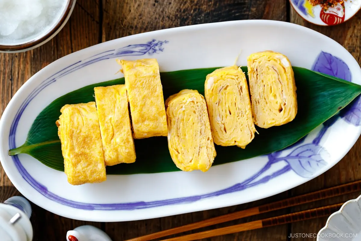 Several slices of tamagoyaki are neatly arranged on a green leaf atop a decorative white plate. The golden-yellow rolled omelet pieces showcase delicate, thin layers and are accompanied by side dishes and chopsticks on a wooden surface.