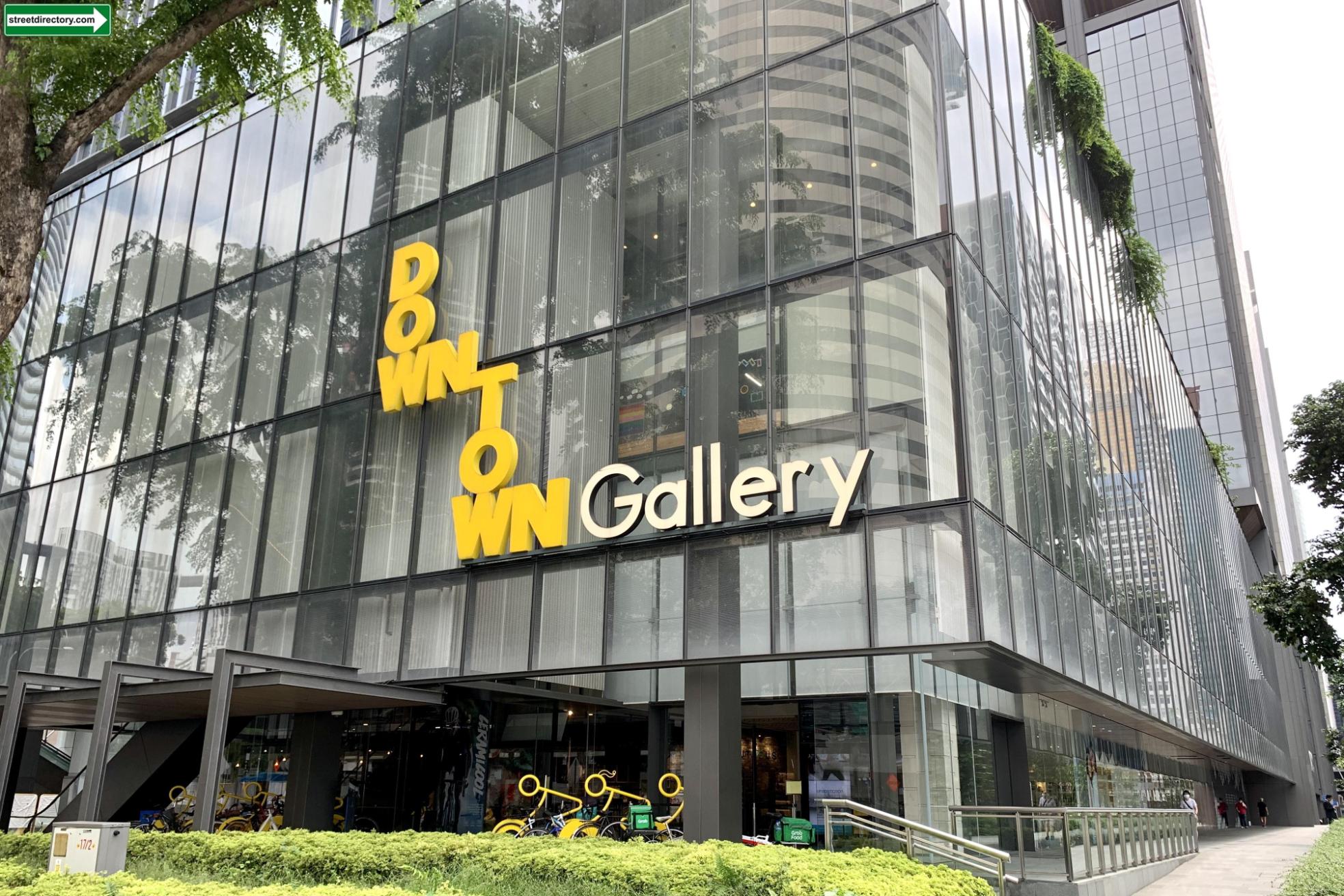 The image depicts the exterior of "Downtown Gallery," a modern multi-story building featuring a sleek glass facade and vibrant yellow signage. The glass reflects surrounding skyscrapers and greenery, while the ground level shows a manicured hedge and a row of parked bicycles.