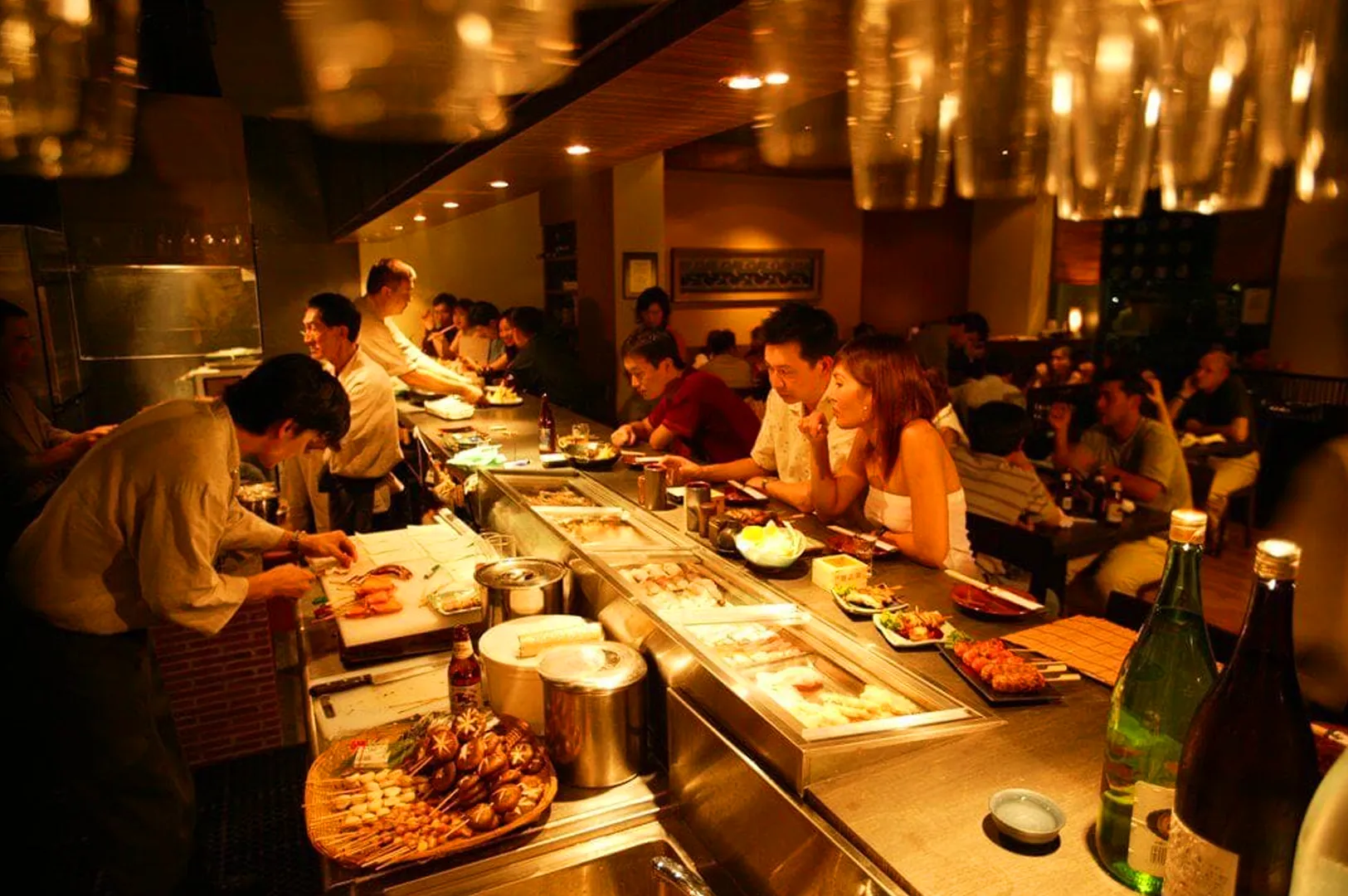 yakitori counter Singapore izakaya chef preparing skewers with diners seated at bar warm lighting Japanese dining atmosphere
