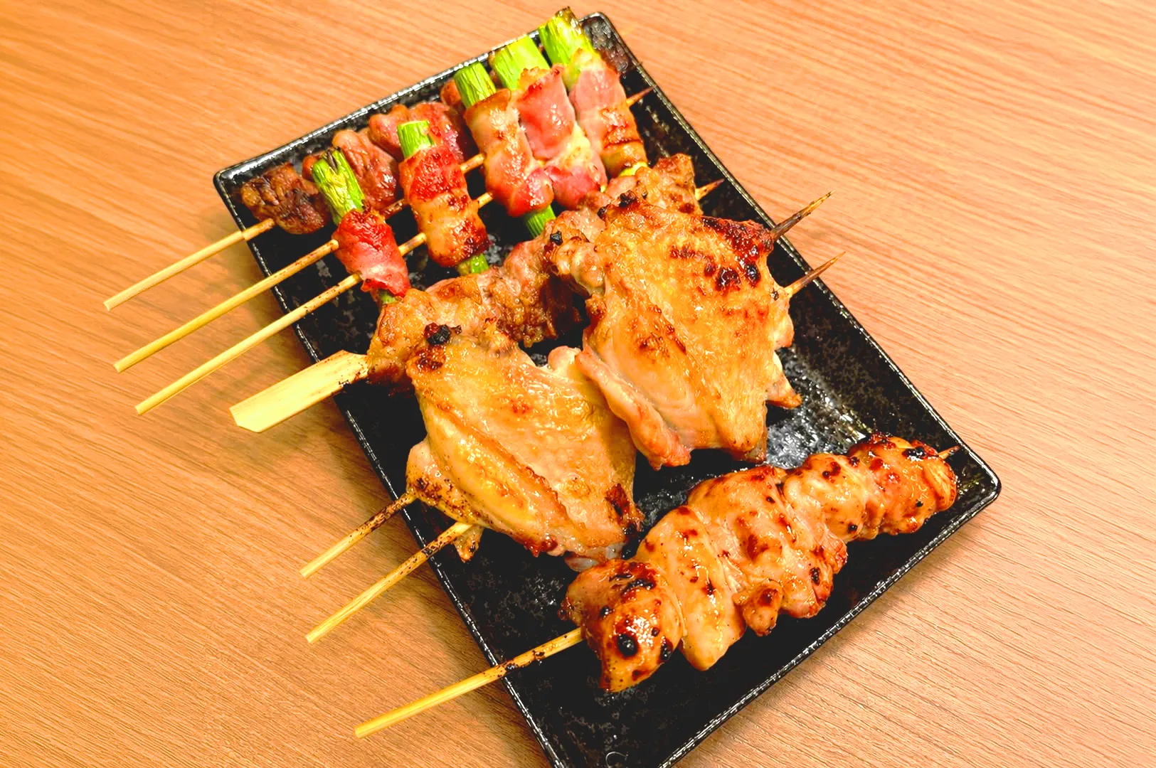 Grilled yakitori skewers with chicken thigh and bacon-wrapped vegetables on a black plate, Japanese izakaya food close-up, Singapore dining scene