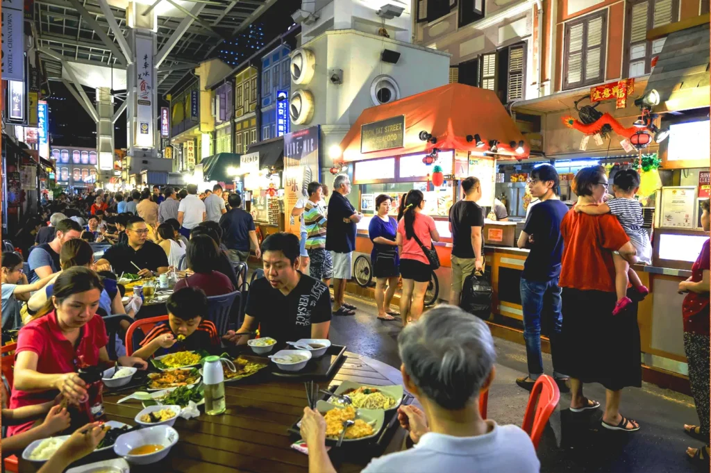 Busy Singapore night street food scene with outdoor dining, hawker stalls, and people eating in a lively evening atmosphere