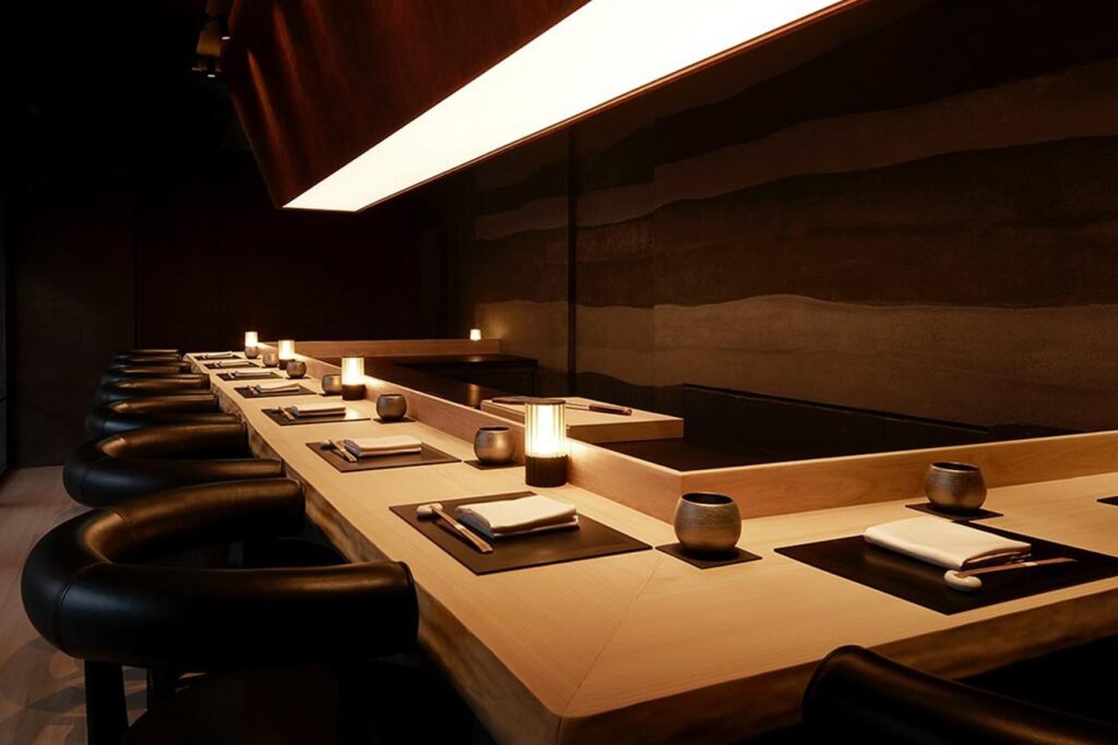 This image showcases a sophisticated, modern sushi counter featuring a sleek light wood surface, minimalist place settings, and curved black leather chairs. The atmosphere is intimate and moody, characterized by warm ambient lighting from small table lamps and a striking, angular overhead light fixture.