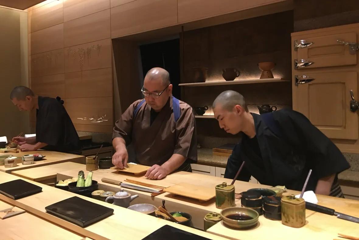 Three chefs are meticulously preparing food behind a light wood counter in an upscale, minimalist Japanese kitchen. The scene is characterized by warm lighting, traditional dark-colored uniforms, and various ceramic vessels and culinary tools organized across the workspace.