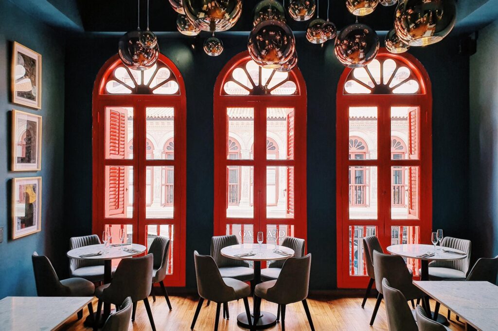 This stylish dining room features three vibrant red arched windows with traditional shutters that provide a view of an adjacent historic building. The interior is accented by a cluster of metallic globe pendant lights hanging from a dark ceiling, complemented by marble-topped tables and grey upholstered chairs.