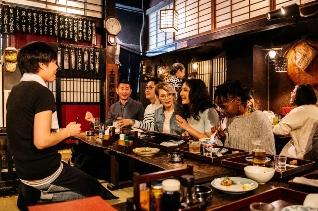 A group of people are seated at a long wooden table in a dimly lit restaurant, enjoying food and drinks. A server is standing and interacting with the guests.