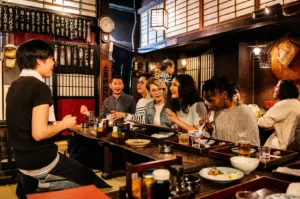 A group of people are seated at a long wooden table in a dimly lit restaurant, enjoying food and drinks. A server is standing and interacting with the guests.