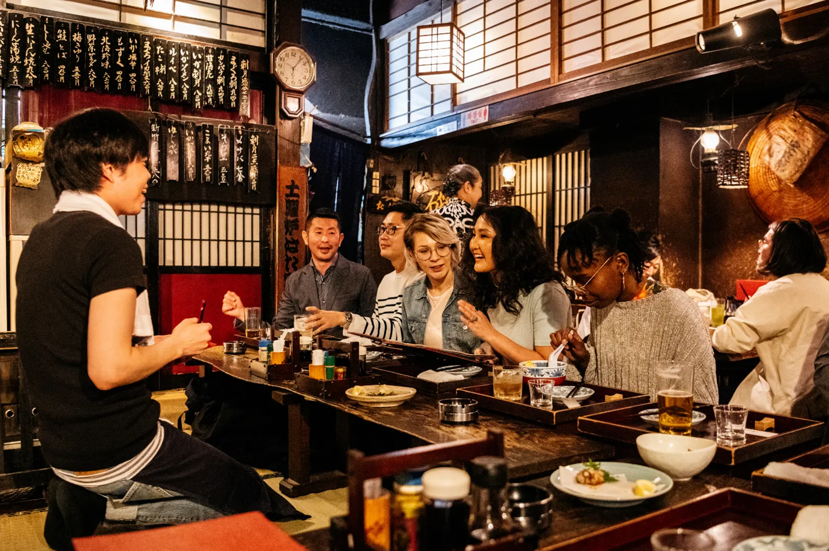 A group of people are seated at a long wooden table in a dimly lit restaurant, enjoying food and drinks. A server is standing and interacting with the guests.