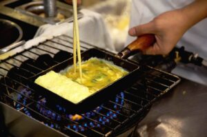 Japanese chef cooking tamagoyaki in rectangular pan at izakaya kitchen close up