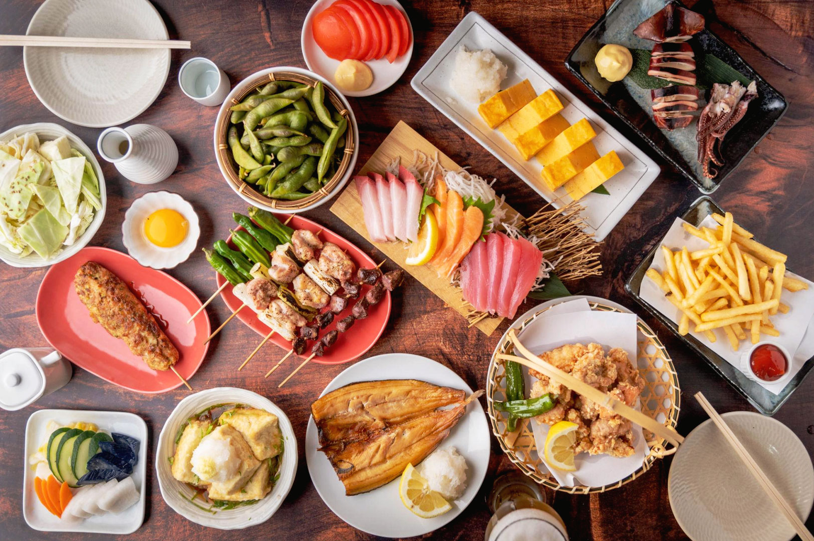 Overhead view of a crowded izakaya table filled with yakitori skewers, sashimi, karaage, tamagoyaki, grilled fish, edamame, and Japanese sharing dishes on small plates