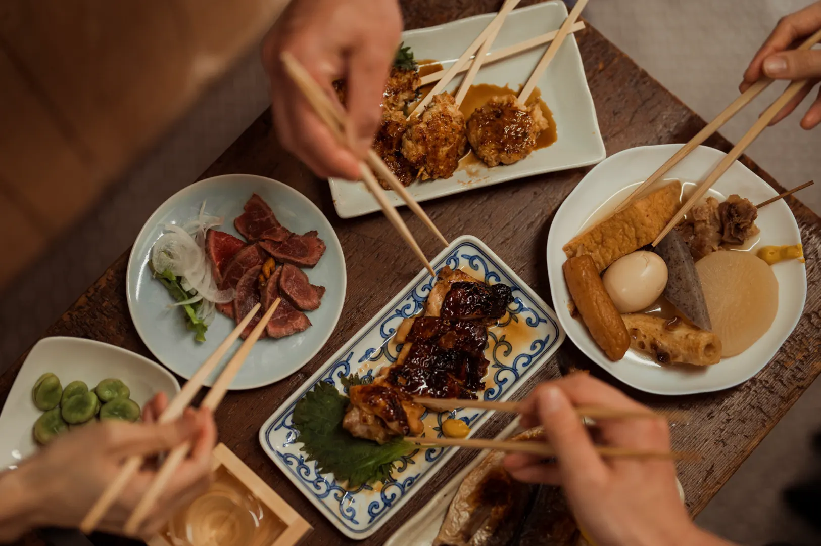 Several plates of food, including oden, grilled fish, and sliced meat, are arranged on a wooden table, with people using chopsticks to eat.