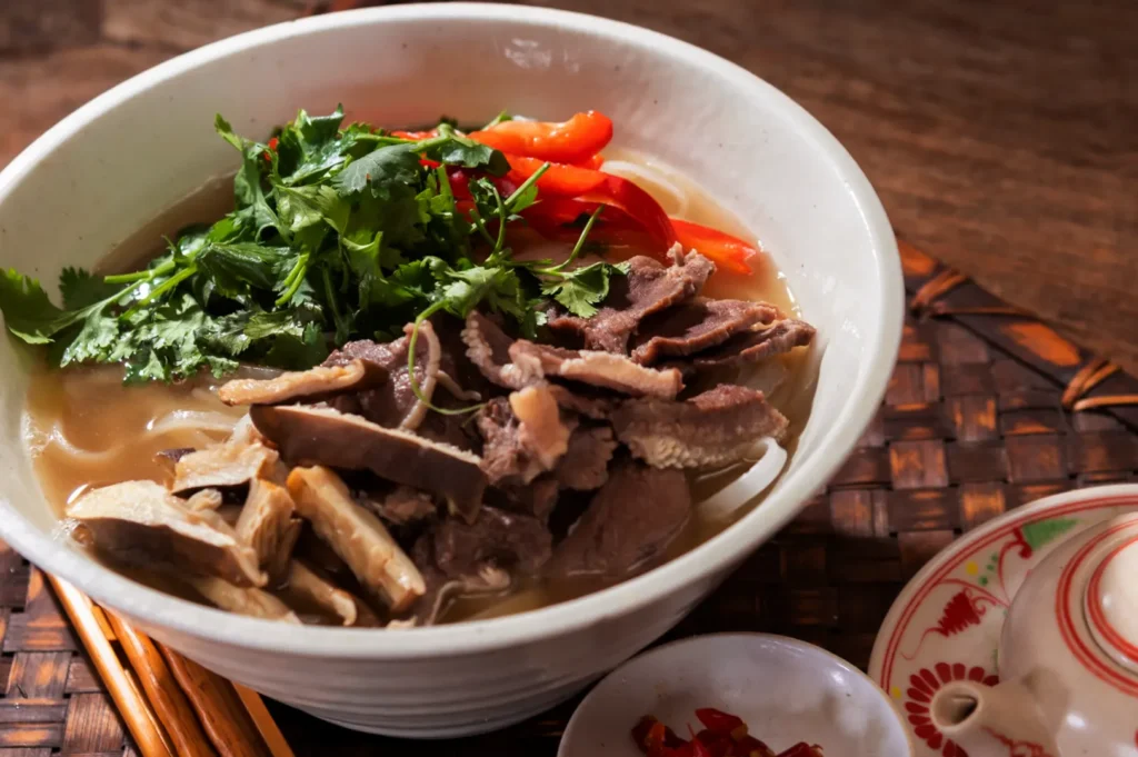 A bowl of pho with offal beef slices, noodles, cilantro, and red pepper slices, served with chopsticks and a small dish of chopped chili peppers.