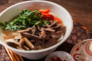 A bowl of pho with offal beef slices, noodles, cilantro, and red pepper slices, served with chopsticks and a small dish of chopped chili peppers.