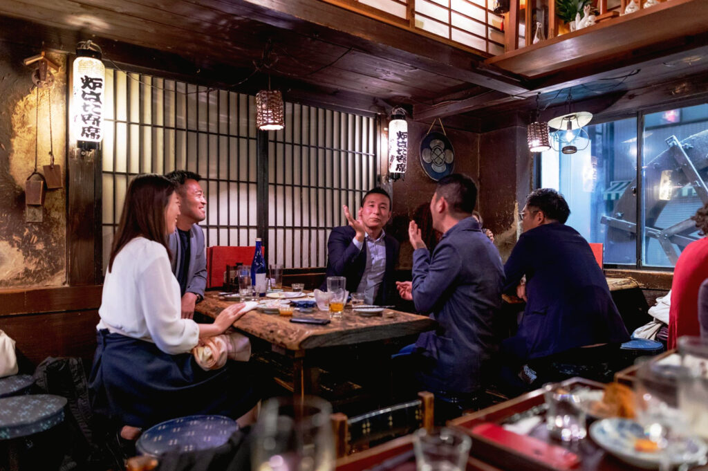 Busy Japanese izakaya with a small wooden table shared by office workers drinking sake and chatting closely in a warm traditional restaurant setting