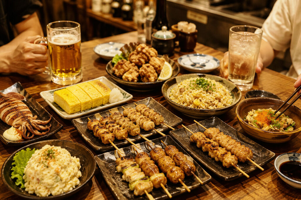 Japanese izakaya table spread with grilled squid, tamagoyaki, chicken skewers, karaage, fried rice, and potato salad in a cozy restaurant setting