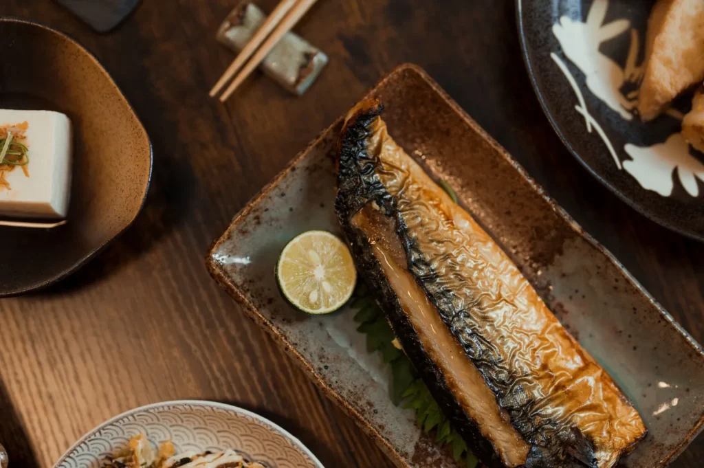 A piece of grilled fish with lime, a cube of tofu with toppings, fried food, and a bowl of noodles or salad.