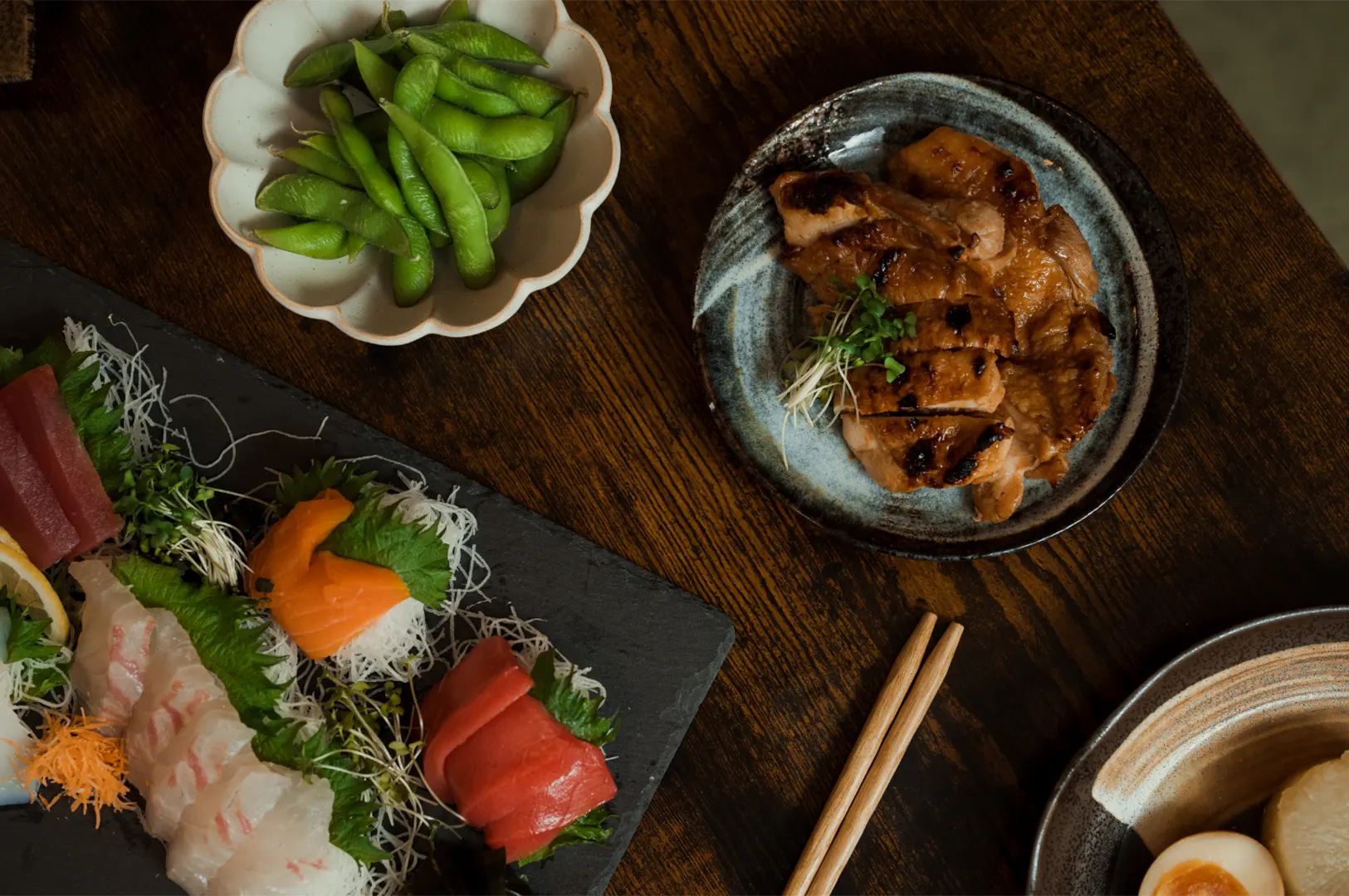 A spread of Japanese food including assorted sashimi, grilled meat, edamame, and a dish with a boiled egg and radish.