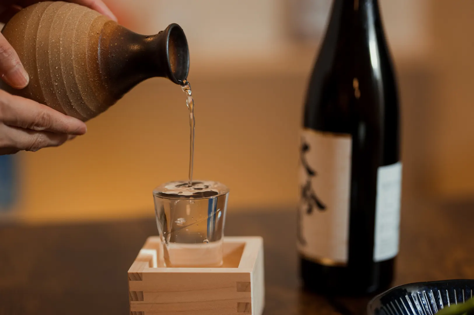 A hand pours sake from a ceramic carafe into a small glass resting in a wooden sake box. A bottle of sake and a plate of sashimi are on the table.