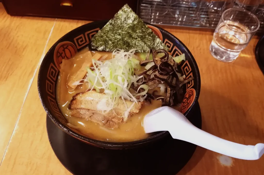 A bowl of ramen with pork, green onions, seaweed, and bamboo shoots, served with a white spoon.