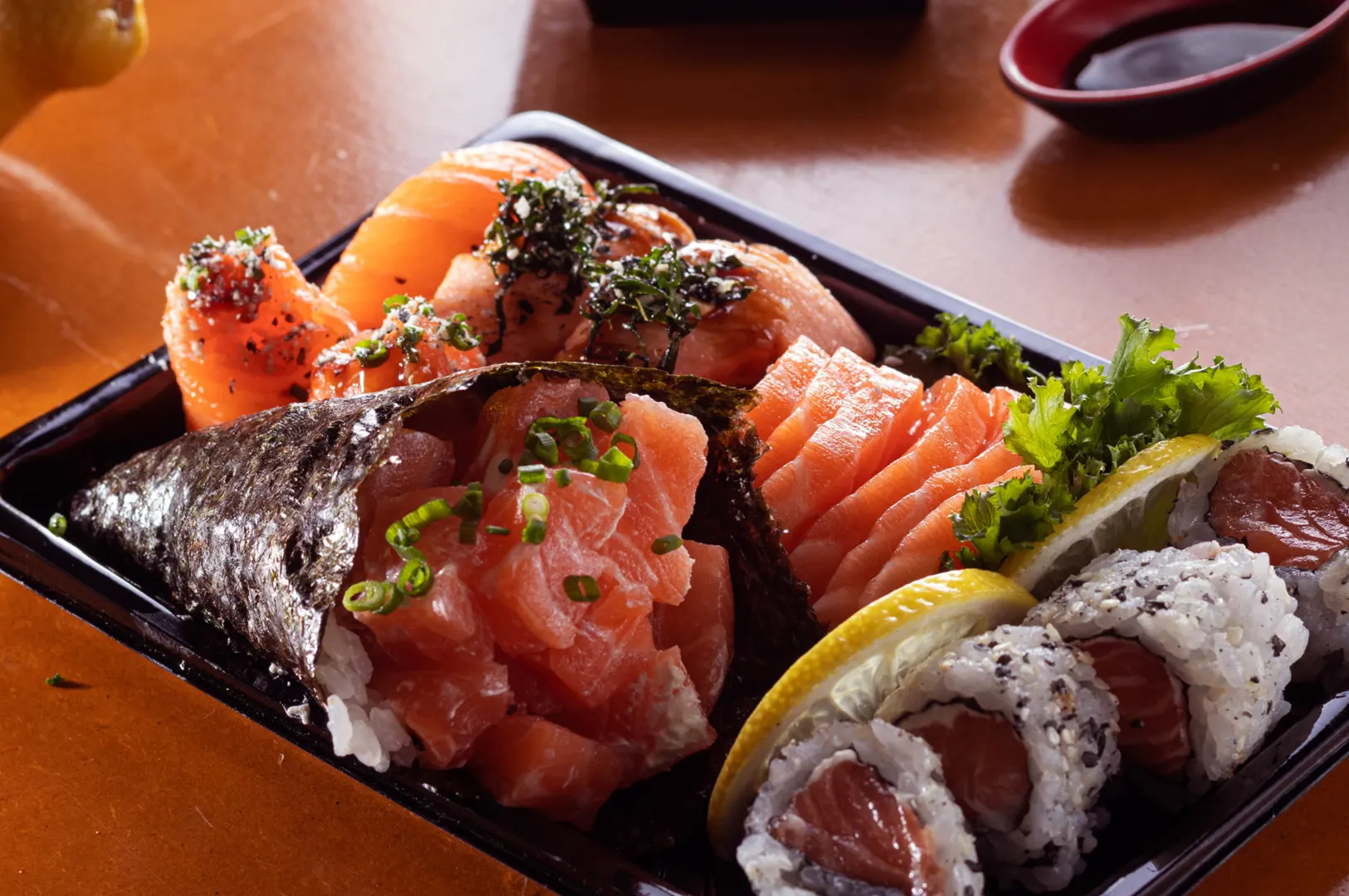A black tray filled with assorted sushi and sashimi, including salmon nigiri, tuna hand roll, salmon sashimi, and California rolls garnished with lemon slices and lettuce.