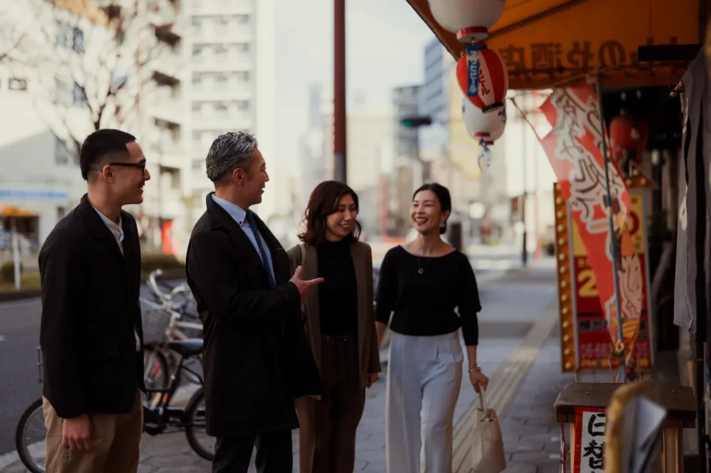 Four people are walking on a sidewalk next to a street, with buildings in the background. A store front with lanterns and signs is on the right.