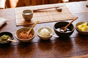 A wooden table with small bowls of Japanese condiments, including yuzu kosho, shichimi, grated daikon, tare, and lemon wedges.