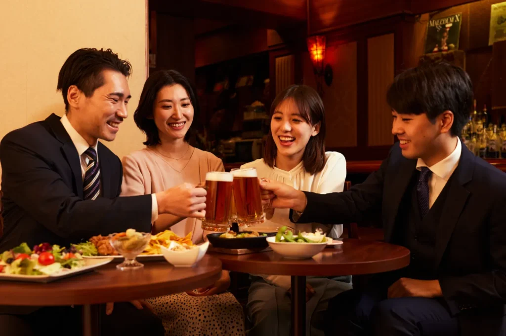 Four people toast beer at a warmly lit izakaya table with plates of food.