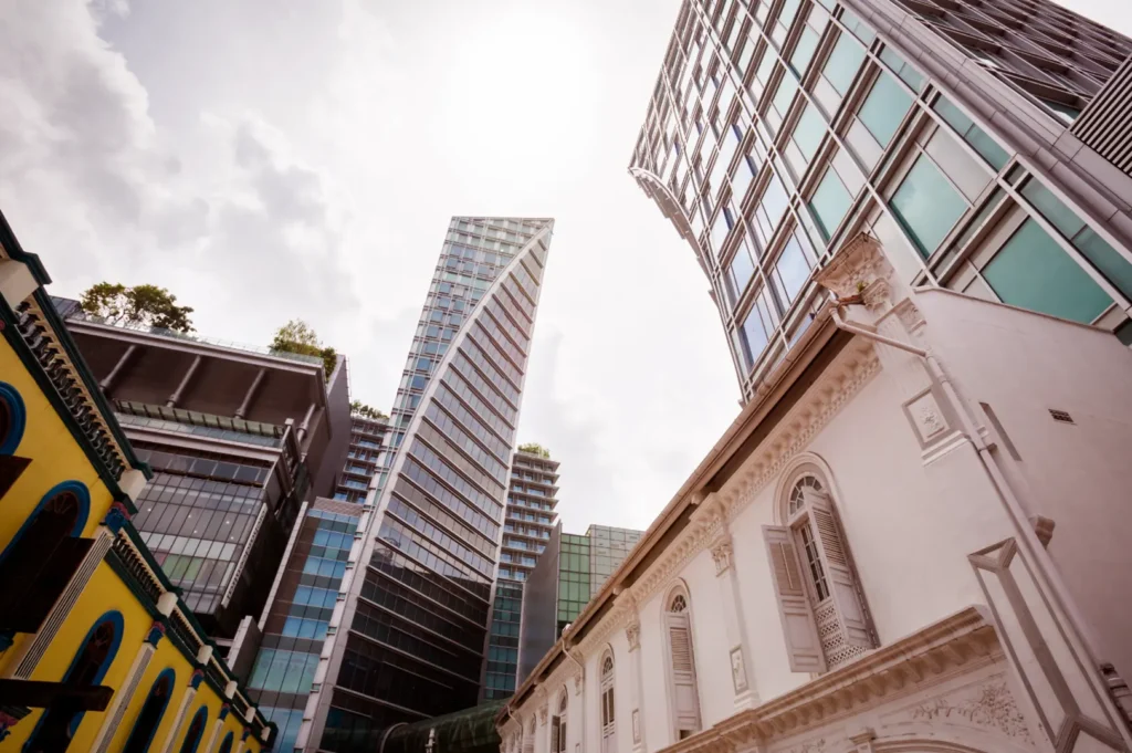 Low-angle view of modern glass high-rise towers above white heritage building with classical architecture, cloudy sky.