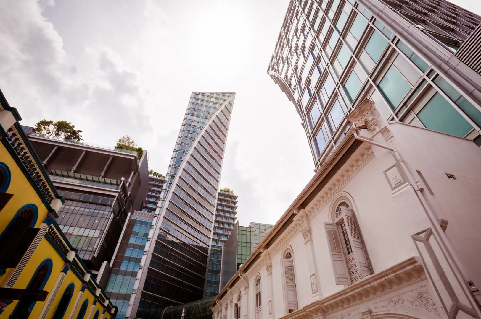 Low-angle view of modern glass high-rise towers above white heritage building with classical architecture, cloudy sky.
