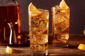 Two whisky highballs with ice and lemon wedges on wooden table.