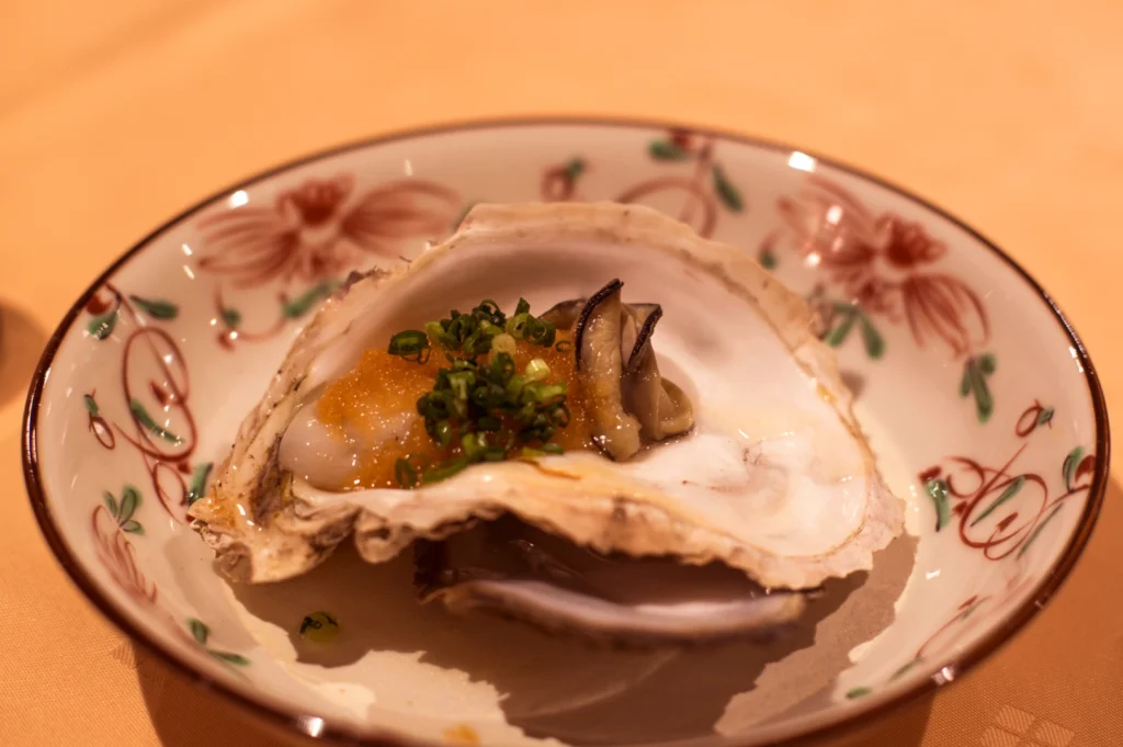 A single oyster served in its shell on a decorative plate, topped with a dollop of orange roe and chopped green onions.