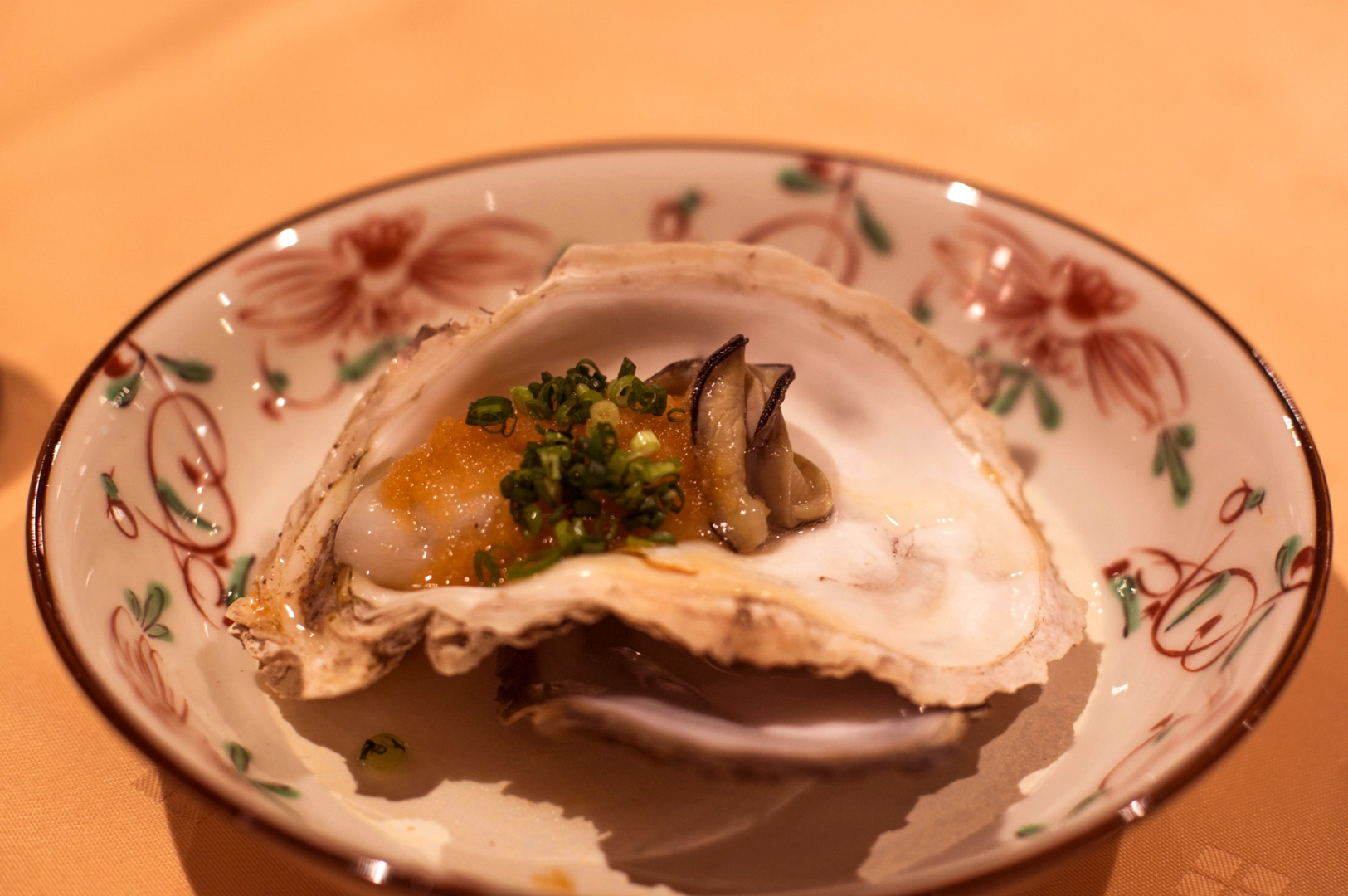 A single oyster served in its shell on a decorative plate, topped with a dollop of orange roe and chopped green onions.