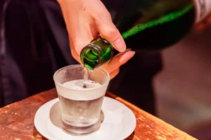 A hand pours liquid from a green bottle into a frosted glass on a saucer.