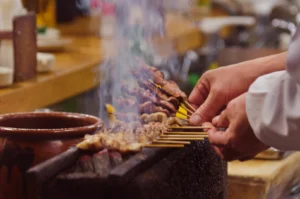 A person grilling skewers of meat over coals, with smoke rising.