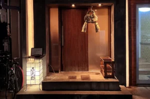 A warmly lit wooden entrance with a hanging rope ornament, glowing lantern, and a bicycle at the side.