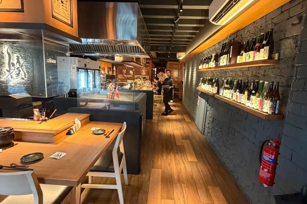 A long robata-style restaurant interior with a counter, exposed grill, and shelves lined with sake bottles.