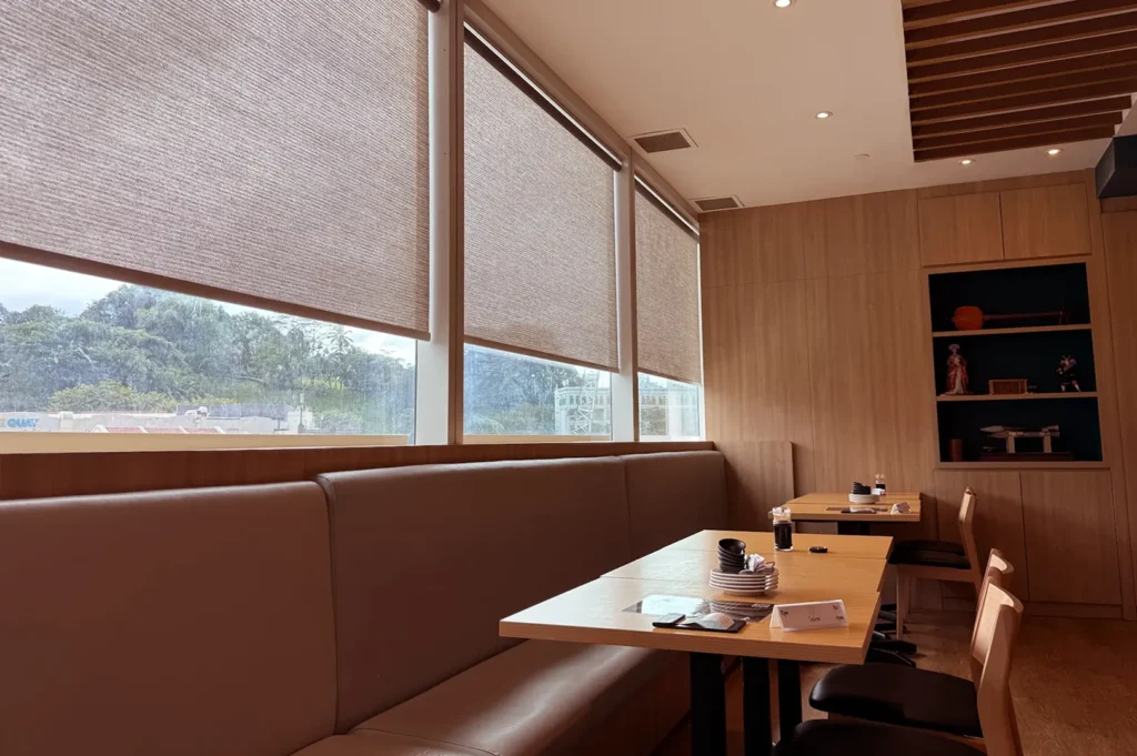 Quiet restaurant interior with booth seating, wooden tables, large window shades, and a warm wood finish.