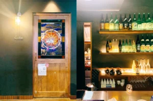 Apollon entrance with stained glass door panel and backlit sake bottle shelves on dark green wall.