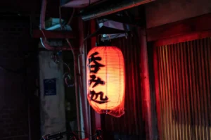 A glowing red izakaya lantern hangs outside a dim wooden storefront at night.