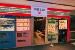 Ume San 100 storefront with bright red signage, an open entrance, and drink vending machine display fridges on both sides.