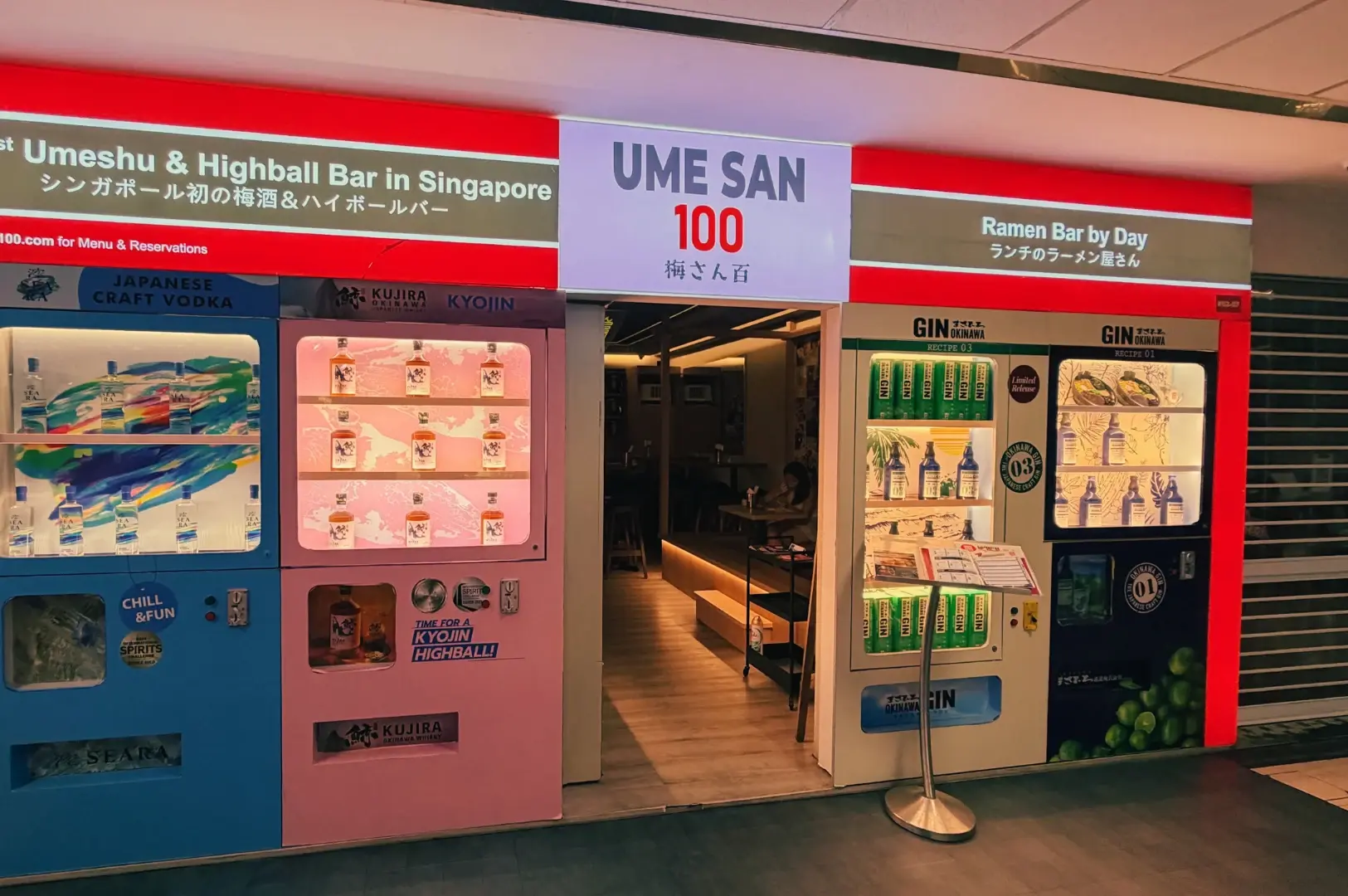 Ume San 100 storefront with bright red signage, an open entrance, and drink vending machine display fridges on both sides.