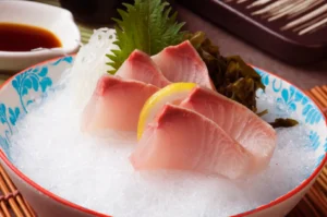 Sliced raw fish garnished with lemon wedges and leafy greens served in a blue-and-red bowl filled with ice, with a small dish of dipping sauce and chopsticks nearby.