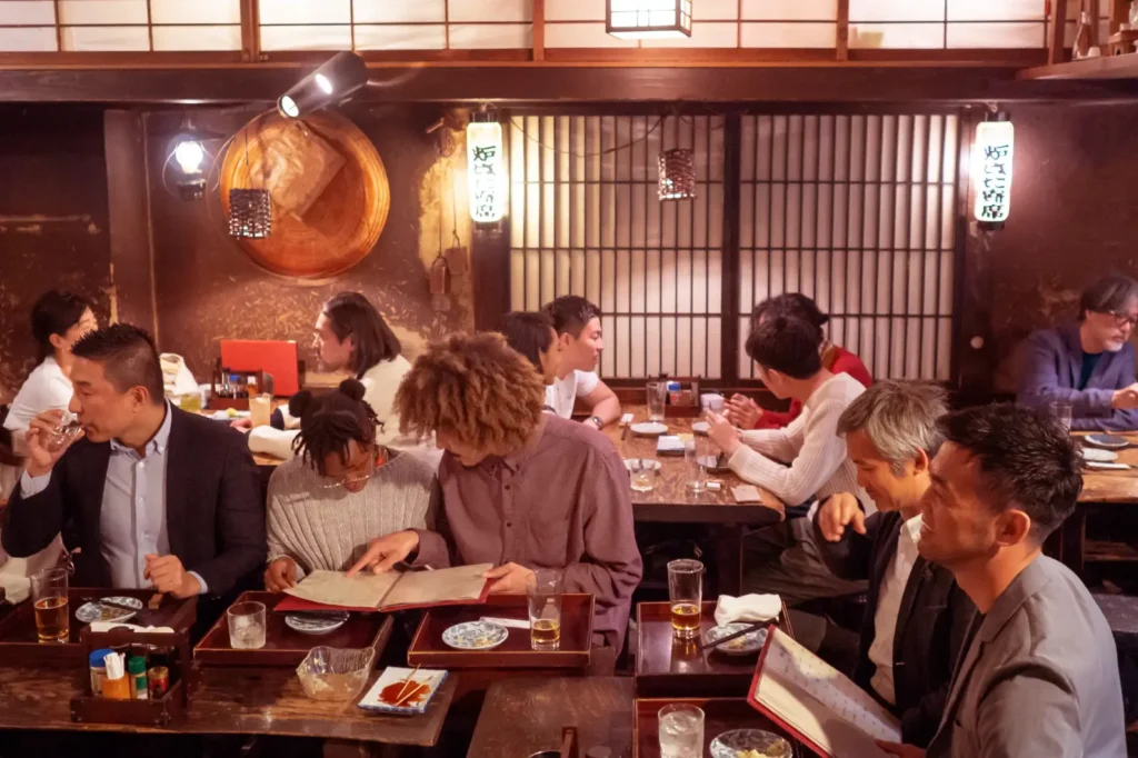 Busy Japanese izakaya dining room with people seated at dark wooden tables eating and drinking, warm lantern lights, and a barred back wall with hanging menu signs.