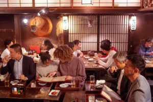 Busy Japanese izakaya dining room with people seated at dark wooden tables eating and drinking, warm lantern lights, and a barred back wall with hanging menu signs.