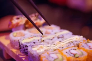 Close-up of sushi rolls on a wooden serving tray, with chopsticks resting over the pieces and visible fish, rice, and orange garnish in soft focus background.