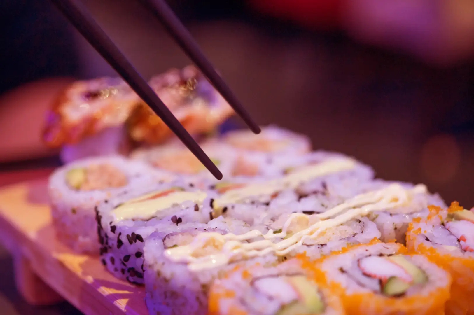 Close-up of sushi rolls on a wooden serving tray, with chopsticks resting over the pieces and visible fish, rice, and orange garnish in soft focus background.