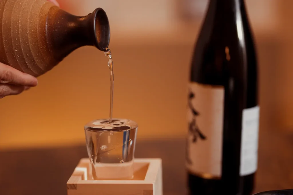 A hand pours sake liquor from a bottle into a small glass on a wooden crate, with another dark bottle at the back.