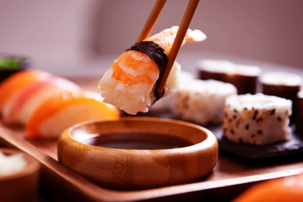 Close-up of sushi on a wooden tray: chopsticks hold a piece of nigiri with salmon and rice, with rolls and maki in the background.