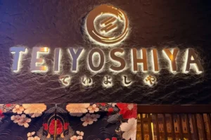 Illuminated TEIYOSHIYA restaurant sign above a floral Japanese-style entrance.