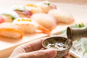 A hand pours a dark sauce from a small ceramic spoon into a tiny bowl held over a tray of assorted bread rolls and shrimp, with lettuce and greens in the background.