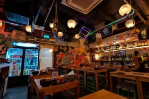 Warmly lit Yorimichi Yakitori Izakaya dining room with wooden tables, hanging lanterns, Japanese artwork, a mural, and shelves of drinks.